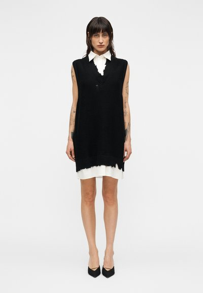 Robe noire en maille sans manches avec un profond décolleté en V et un ourlet effiloché, portée sur une chemise blanche à col. Assortie à des talons noirs pointus.
