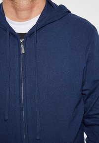 Marinefarbener Zip-Hoodie aus strukturiertem Material, mit Kapuze, Kordeln und einem silbernen Reißverschluss. Nahaufnahme des Oberkörpers.
