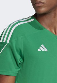 Jersey atlético verde con cuello en V, tres franjas blancas en cada hombro y un acento texturizado en la parte superior de la manga. Logo blanco exhibido.