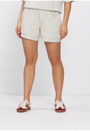 Vrouw draagt lichtgrijze casual korte broek en witte sneakers met rode accenten, staand tegen een effen witte achtergrond.