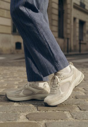 Person wearing white athletic sneakers and blue pinstriped pants, walking on cobblestone street in urban setting.