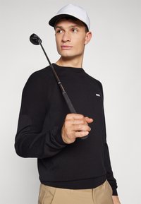 Schwarzer Pullover mit strukturiertem Design, ausgestattet mit einem kleinen weißen Logo. Das Modell hält einen Golfschläger und trägt eine weiße Mütze. Beige Hosen runden den Look ab.