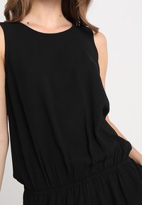 Czarna sukienka bez rękawów z okrągłym dekoltem, marszczonym pasem i miękkim, lejącym materiałem, charakteryzująca się luźnym krojem i gładką fakturą.