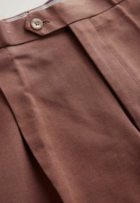 Brown woven fabric with a visible texture, featuring a front pleat and a button detail at the waistband.