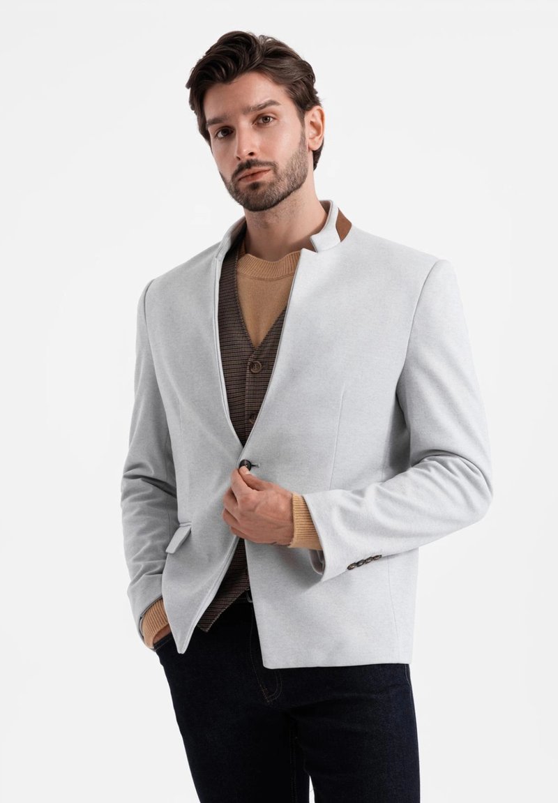 Blazer grigio chiaro con colletto alla mandarino, dotato di due tasche frontali e un maglione marrone texturizzato indossato sotto. Tessuto morbido.