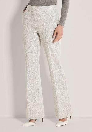 Femme portant un pantalon blanc taille haute à motif paisley, jambes évasées et talons blancs pointus, debout avec une main dans la poche.
