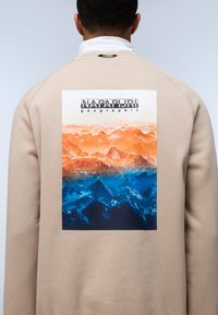 Sudadera beige con un gran gráfico de un paisaje montañoso colorido en naranja y azul, con el texto "NAPAPIJRI geographic" sobre la imagen.