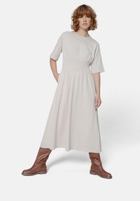 Vestito beige chiaro, alla lunghezza del ginocchio, con maniche corte, vita aderente e gonna arricciata. Abbinato a stivali marroni al ginocchio. Texture liscia.