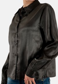 Camicia nera in raso con bottoni, collo a risvolto, maniche lunghe e polsini elasticizzati. Il tessuto ha una texture liscia e lucida.