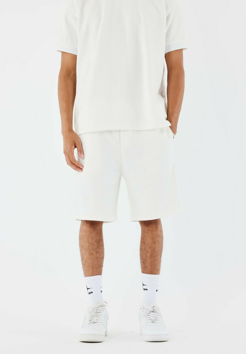 Camisa de punto blanca de manga corta combinada con pantalones cortos blancos a juego. El conjunto presenta un corte relajado y texturas sutiles. Zapatillas blancas completan el look.