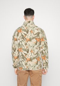 Giacca in pile beige con un motivo floreale arancione e verde, dotata di un colletto alto e una vestibilità rilassata, mostrata di spalle.