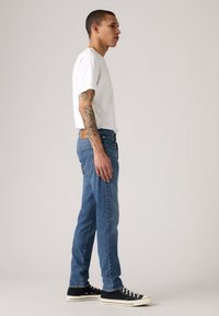 Fatos de ganga azul com uma cintura escura, corte ajustado, combinados com uma t-shirt branca de decote redondo e ténis pretos de cano alto.