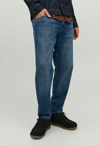 Jeans in denim blu scuro con taglio dritto, caratterizzati da leggere scoloriture e dettagli di cucitura. Indossati con scarpe in suede nere e una giacca scura.