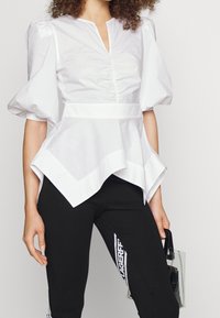 Blusa peplum bianca con maniche a sbuffo, caratterizzata da una scollatura a V e dettaglio raccolto nella parte frontale, abbinata a leggings neri con logo bianco.