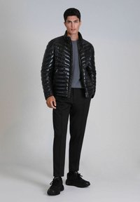 Zwarte pufferjack met een glanzende afwerking, ritssluiting aan de voorkant en eenstaande kraag, gecombineerd met een grijs shirt en zwarte op maat gemaakte broek. Zwarte sneakers.
