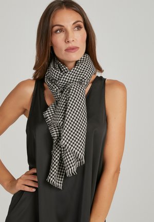 Femme portant un haut noir sans manches et une écharpe à carreaux noirs et blancs nouée autour du cou, regardant légèrement vers le haut et sur le côté.