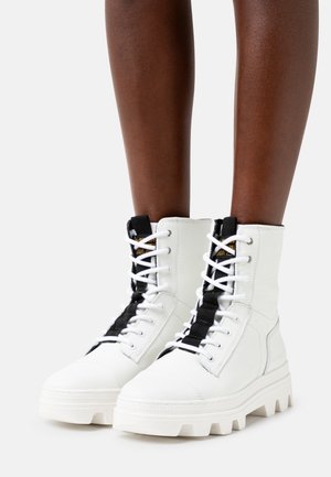 Bottes blanches à lacets montantes avec des sangles centrales noires portées sur des jambes à la peau foncée, sur un fond clair uni.