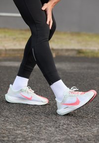Lichte Nike sneakers met een bovenwerk van mesh, voorzien van mintgroene en felroze accenten, aangevuld met witte veters en sokken, gedragen met zwarte leggings.