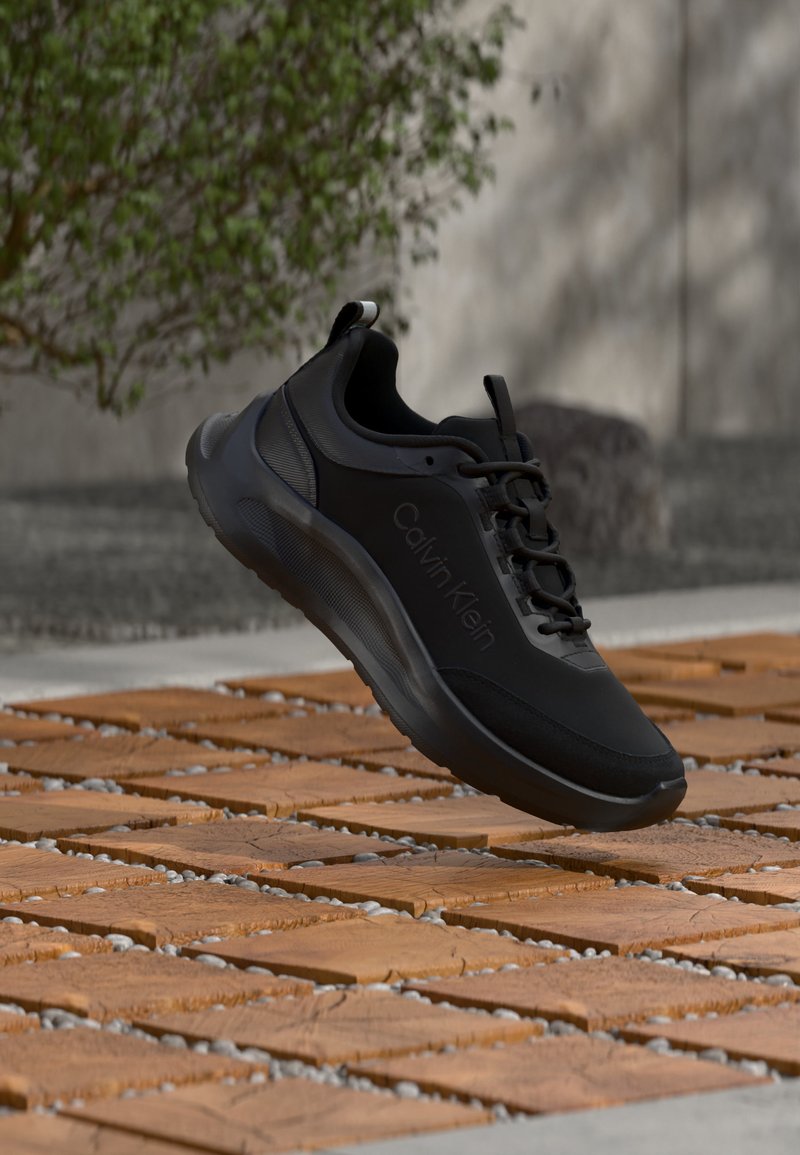 Baskets noires Calvin Klein à semelle épaisse flottant au-dessus de dalles en bois avec de petits cailloux, mur flou et végétation en arrière-plan.