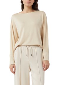Beżowa bluzka z długim rękawem o szerokim dekolcie, luźnym kroju i miękkiej fakturze, zestawiona z jasnymi spodniami na sznurek.