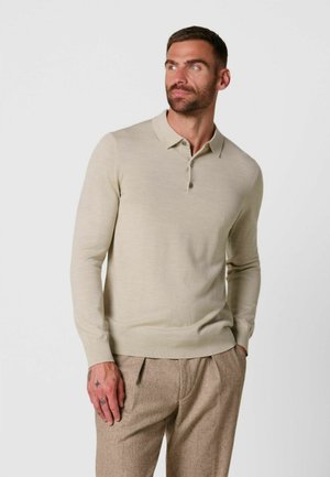 Man met tatoeages die een beige lange mouwen poloshirt en lichtbruine geplooide broek draagt, kijkt opzij met één hand in zijn zak.