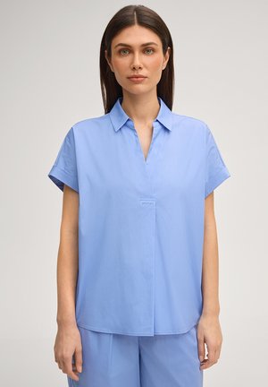 Vrouw die een lichtblauwe blouse met korte mouwen en kraag draagt, en bijpassende broek, staand tegen een effen neutrale achtergrond.