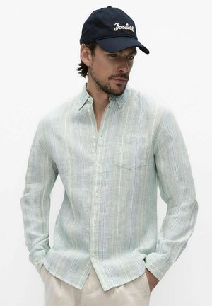 Man draagt een licht gestreept overhemd met lange mouwen en knopen, beige broek en een marineblauwe pet met witte tekst, handen in de zakken, kijkt naar rechts naar beneden.