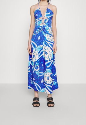 Femme portant une robe à fleurs bleu et blanc avec un col halter, des découpes à l'avant et des sandales noires à volants, debout devant un fond clair uni.