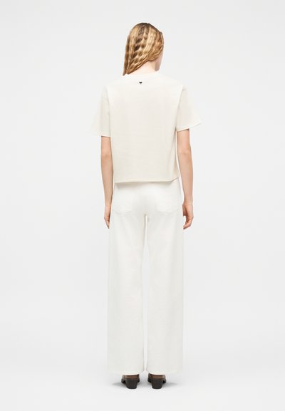 Γυναίκα με μακριά κυματιστά μαλλιά, γυρισμένη με την πλάτη στην κάμερα, φοράει μια λευκή κοντομάνικη μπλούζα και λευκό παντελόνι με φαρδιά μπατζάκια, μαζί με μαύρα παπούτσια.