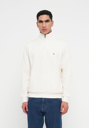 Witte zip-up sweatshirt gemaakt van zachte stof, met een hoge kraag en een klein logo op de borst. Gecombineerd met blauwe denim jeans.