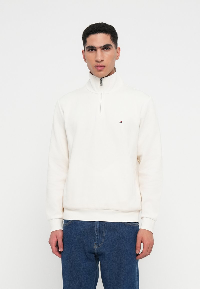 Felpa bianca con zip realizzata in tessuto morbido, dotata di un colletto alto e un piccolo logo sul petto. Abbinata a jeans in denim blu.