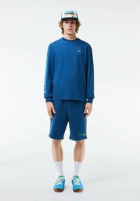 Camisa de manga larga azul con logo verde, pantalones cortos azules a juego y zapatillas azules. Los accesorios incluyen una gorra azul con un logo multicolor.