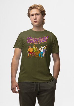 Jonge man met olijfgroene T-shirt met kleurrijke Scooby-Doo-personages en logo, staand met één hand in zijn zak tegen een witte achtergrond.