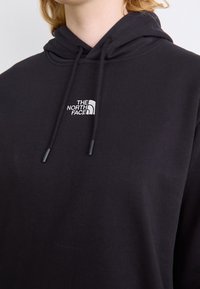 Personne portant un sweat à capuche noir avec cordons et un logo blanc "The North Face" sur la poitrine.