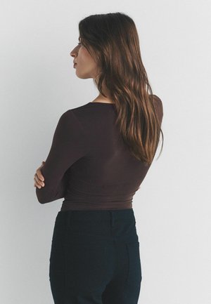 Pikkade pruunide juustega naine tumedas ülaosas ja mustades pükstes, seisab käed risti, vasakule poole, ühevärvse heleda tausta ees.