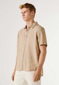Jonge man met kort lichtbruin haar, gekleed in een beige korte mouwen overhemd met knopen en witte broek, tegen een effen achtergrond.
