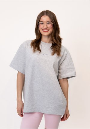 Ung kvinde med langt brunt hår og briller smiler, iført en lys grå oversized T-shirt og blege lyserøde bukser mod en ensfarvet baggrund.