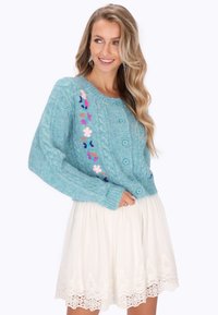 Cárdigan de punto azul claro con patrones de trenzado, bordado floral y cinco botones. Combinado con una falda blanca con encaje.