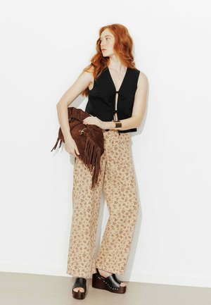 Femme aux cheveux roux portant un haut noir sans manches, un pantalon large beige à motifs floraux, des sandales à plateforme noires, tenant un sac marron à franges, debout contre un mur blanc.