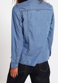 Persona che indossa una camicia di denim blu con piccoli motivi bianchi, polsini abbottonati e jeans scuri, mostrata di spalle.