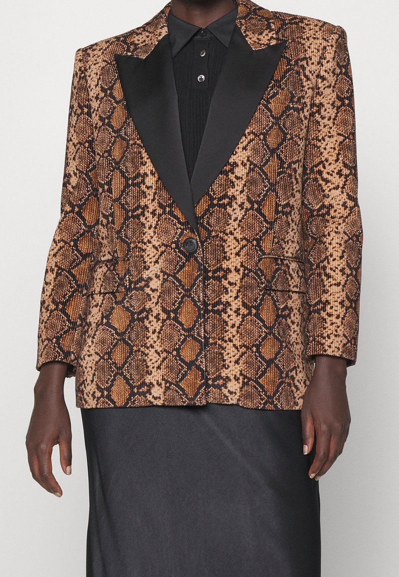 Blazer en peau de serpent avec motifs marron et noir, revers en satin, fermeture par un bouton unique et deux poches latérales. Porté sur une chemise noire texturée.