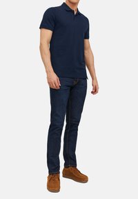 Polo shirt blu navy con colletto, abbinato a jeans blu scuro. Le scarpe sono in suede marrone con un design con lacci e una texture morbida.
