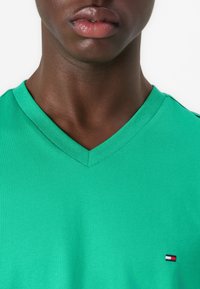 T-shirt vert en V fabriqué en coton doux. Présente un petit logo de marque sur le côté gauche de la poitrine ; manches courtes, design ajusté et texture uniforme.