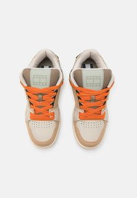 Beige ja tan tennisjalatsid, millel on oranžid paelad, võrgusilma detailid ja logo ploosike keele peal. Ventilatsiooniks perforatsioonidega varbapind.