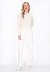 Beżowy sweter w splot warkoczowy z okrągłym dekoltem, noszony z białymi dżinsami w stylu dzwonów i białymi trampkami z czarnymi paskami. Poziome prążkowanie na mankietach.