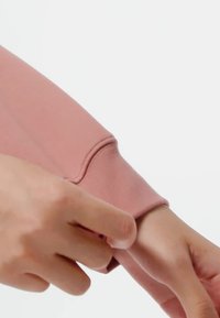 Ruka podešava manžetu od mauve dugih rukava, ističući teksturu tkanine i detalje šivanja.