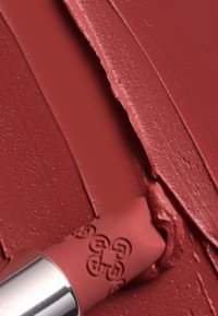 Colore labbra opaco in una tonalità di rosso intenso con una texture liscia. Il tubetto presenta un cappuccio argentato e un logo in rilievo. Campione del prodotto mostrato su una superficie.