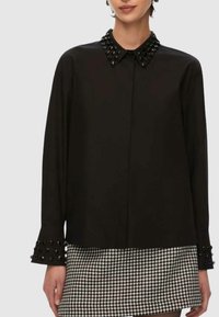 Camicia nera con bottoni, colletto e polsini decorati, dalla texture liscia e maniche lunghe. Indossata sopra una gonna con motivo pied de poule.