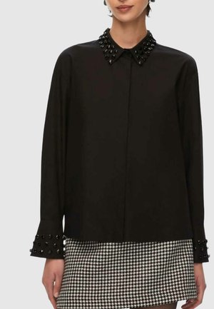 Camicia nera con bottoni, colletto e polsini decorati, dalla texture liscia e maniche lunghe. Indossata sopra una gonna con motivo pied de poule.