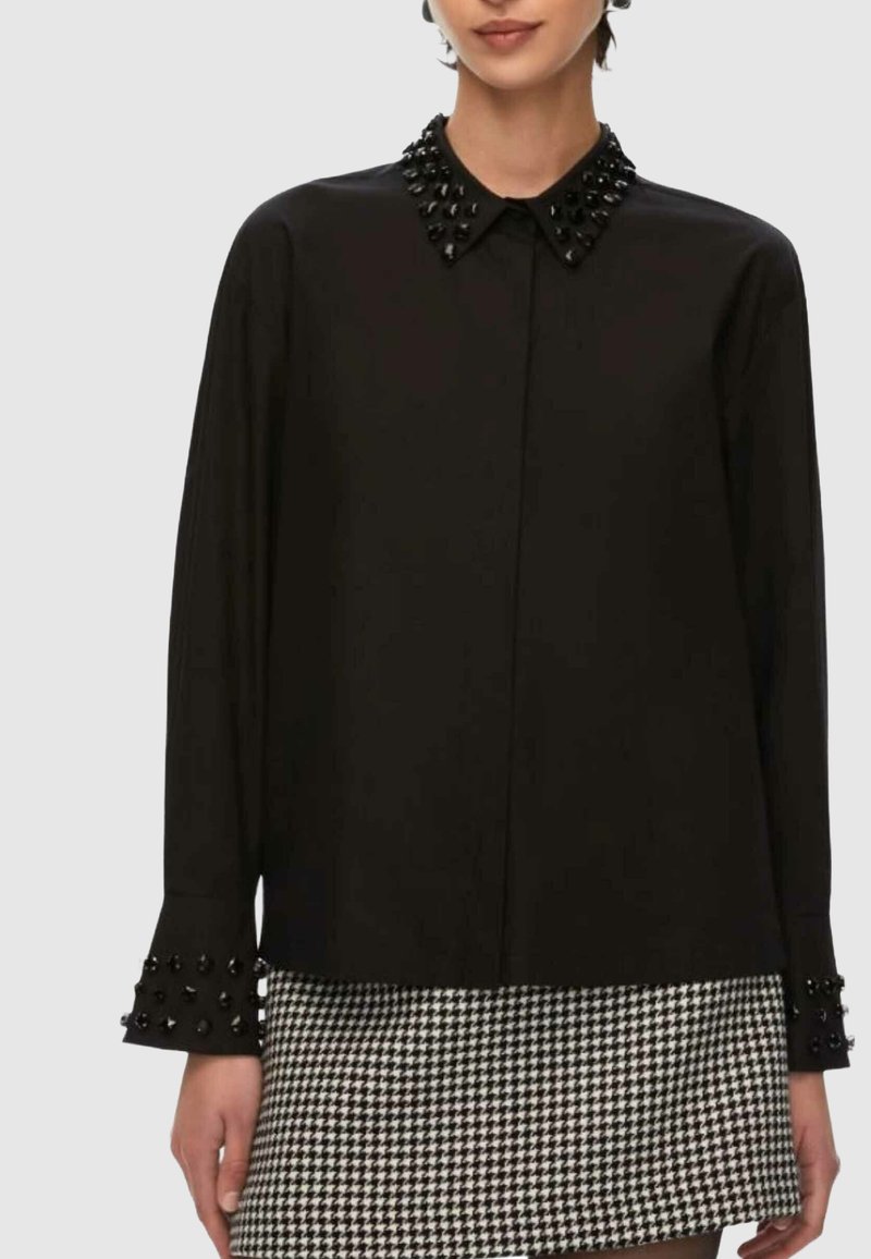 Camicia nera con bottoni, colletto e polsini decorati, dalla texture liscia e maniche lunghe. Indossata sopra una gonna con motivo pied de poule.